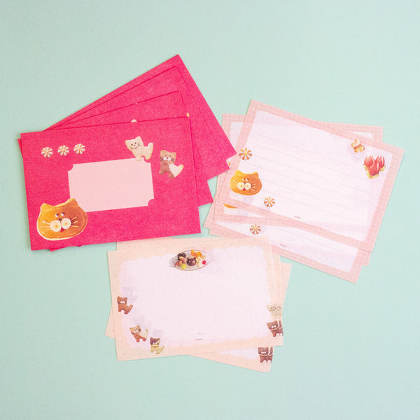 A set of cute stationery items including pink envelopes and lined writing paper sheets decorated with cartoon cat illustrations, arranged on a light mint green background.