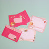 A set of pink stationery items, including envelopes and lined writing paper cards, decorated with cute cartoon food illustrations, arranged on a light mint green background.