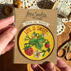 Iron-on patch with 'Life is Sweet' design held by a hand, on a wooden surface with scissors and thread.