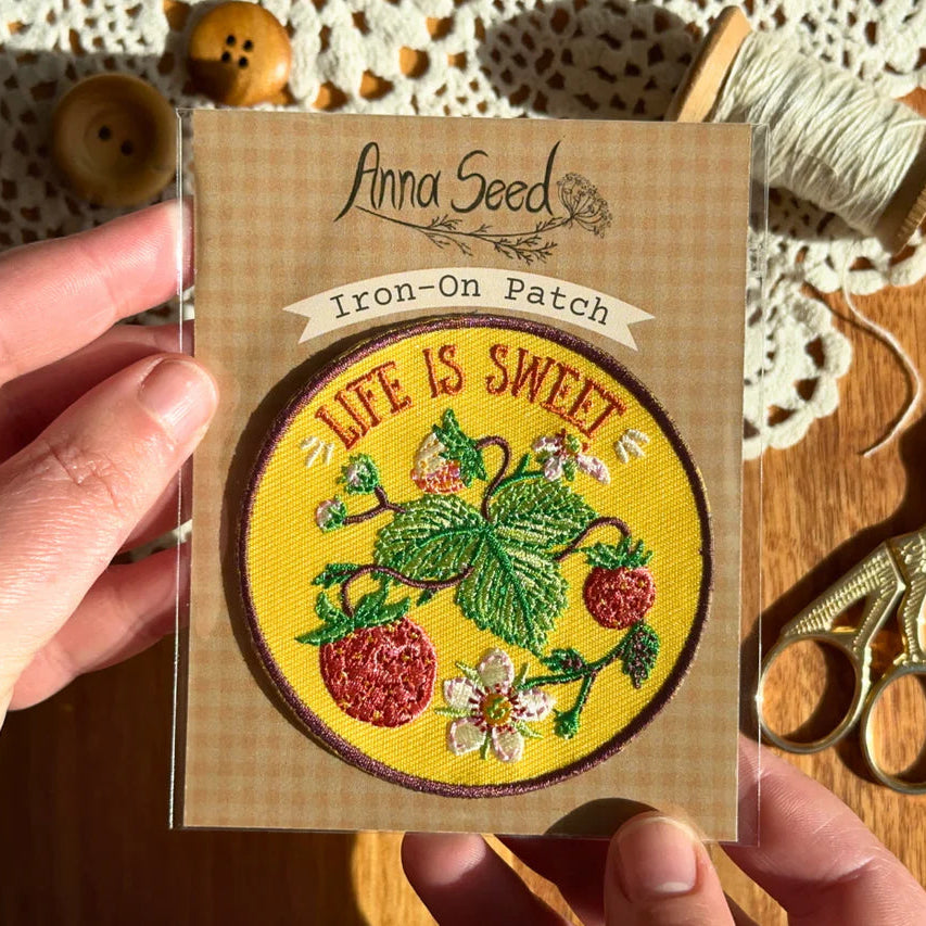 Iron-on patch with 'Life is Sweet' design held by a hand, on a wooden surface with scissors and thread.
