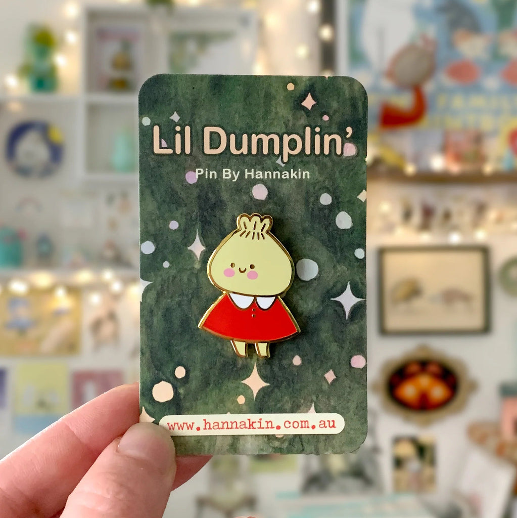 A hand holds a small enamel pin on its backing card. The pin features a cute character with a yellow face and hair wearing a red dress, set against a dark green background with white stars.