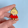A hand holding a small enamel pin shaped like a cartoon character with a yellow round head, pink cheeks, a red dress, and gold outlines, set against a light blue background.