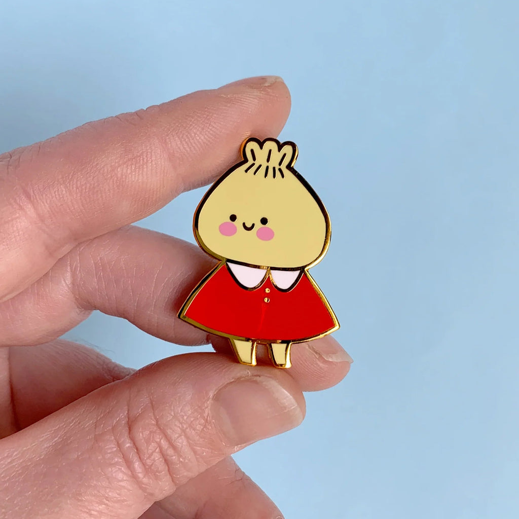 A hand holding a small enamel pin shaped like a cartoon character with a yellow round head, pink cheeks, a red dress, and gold outlines, set against a light blue background.