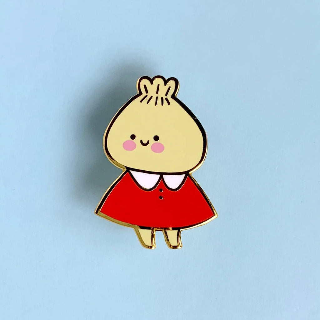 A small enamel pin of a cute cartoon character with a beige head, pink cheeks, a red dress, and a white collar, centered on a light blue background.