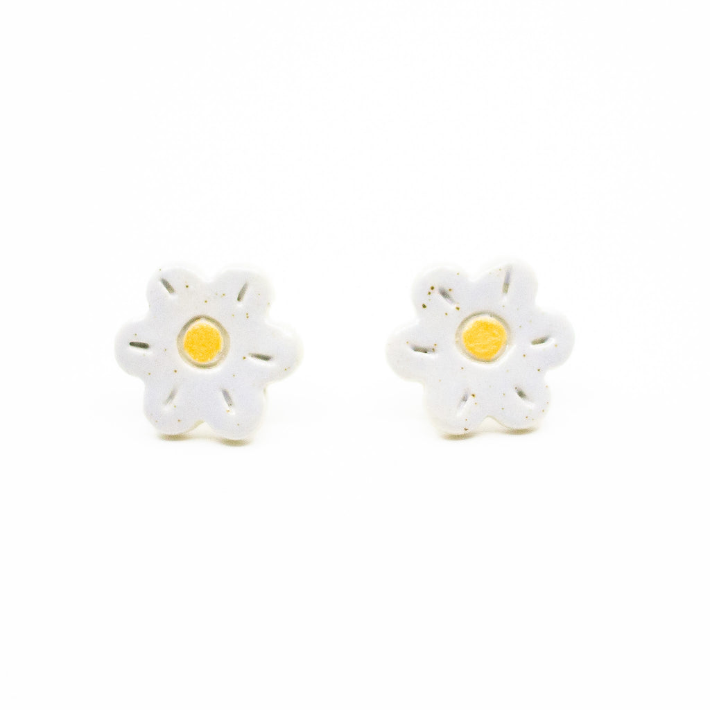 A pair of handmade ceramic stud earrings shaped like white flowers with yellow centres. The earrings have subtle carved details and a glossy finish with natural speckles.