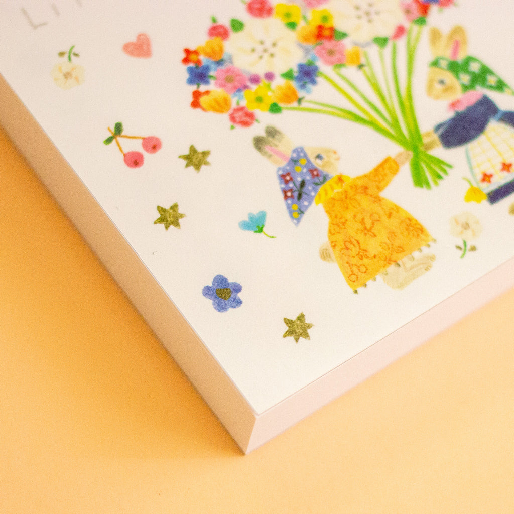 A close-up of a white memo pad cover featuring whimsical illustrated characters, flowers, and small metallic star stickers, set against a warm yellow background.