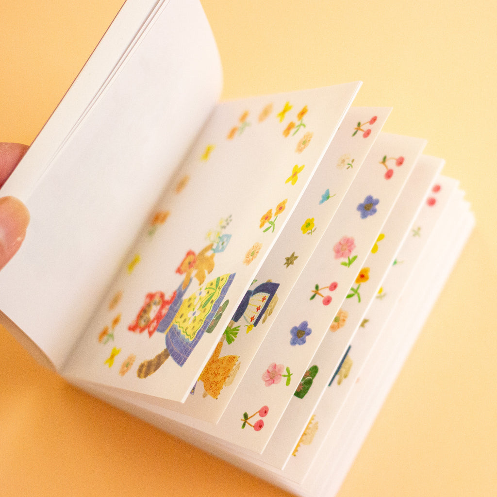 A hand holds an open white notebook  against a warm yellow background. The pages are decorated with small colourful illustrated stickers, including flowers, stars, and whimsical character designs.