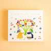 A square memo pad titled 'Little fluffy friends' by Aiko Fukawa, featuring a colourful illustration of two rabbits surrounded by flowers, set against a warm peach background.