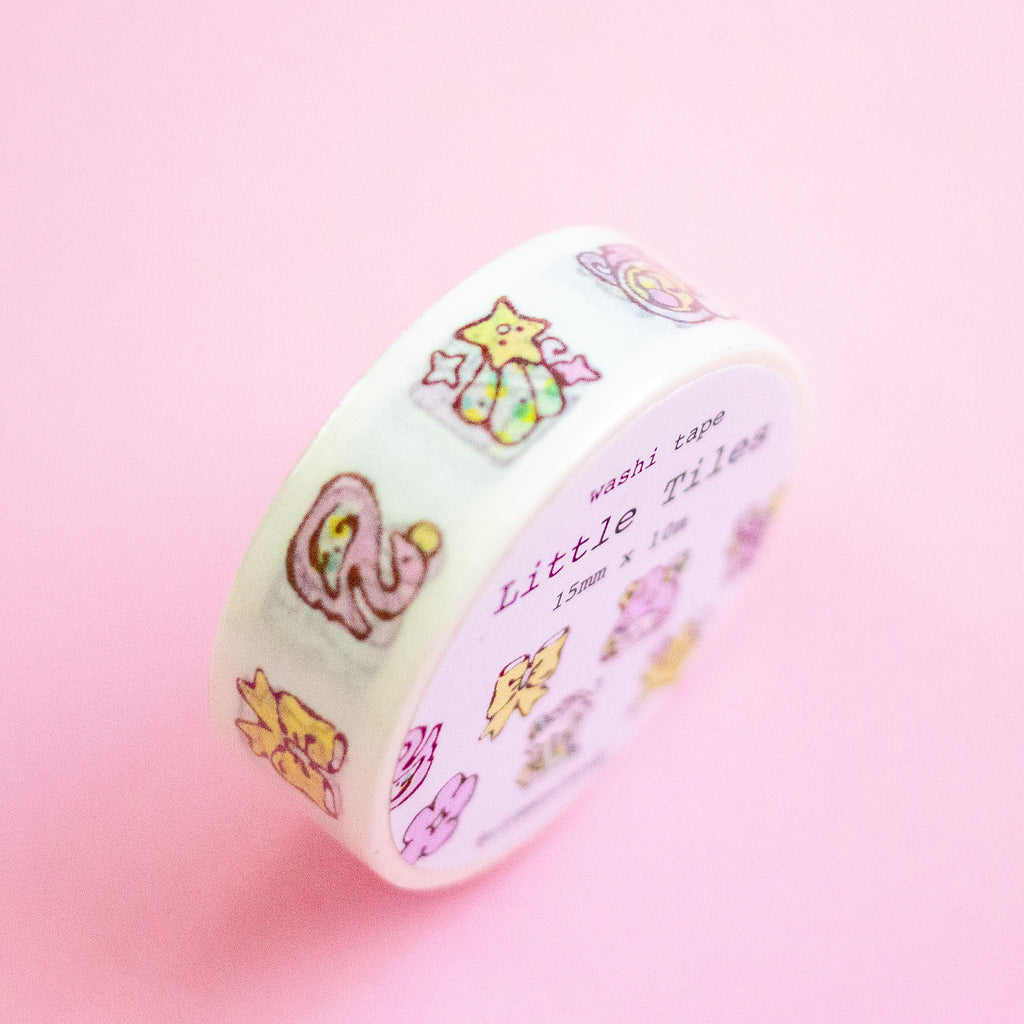 A roll of washi tape with cute pastel illustrations of animals and stars, labeled 'Little Tiles' and 'washi tape', set against a solid pink background.