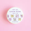 A round container of washi tape labeled 'Little Tiles' by @riomuniandy, featuring cute pastel illustrations of bows, flowers, a cupcake, and stars, set against a solid pink background.