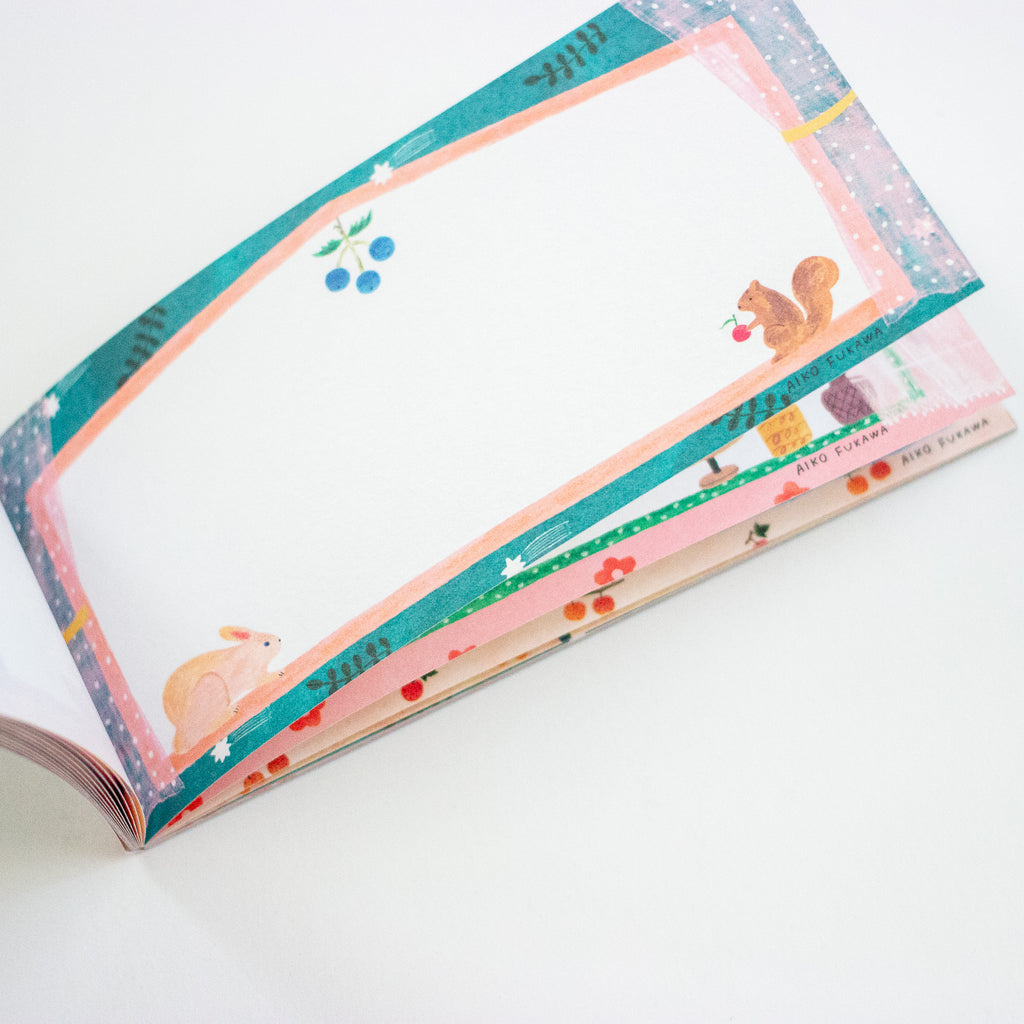 A stack of decorative paper notepads with a whimsical forest theme, featuring illustrated squirrels, a rabbit, berries, and colourful patterned borders on a clean white background.