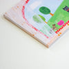 A close-up of a notepad with a whimsical landscape illustration, featuring green trees, a pink sun, and a black cat on a light background.