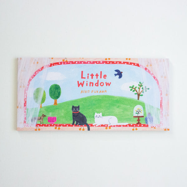A notepad titled 'Little Window' by Aiko Fukawa, featuring a colourful illustration of a green hill, cats, trees, and a bird under a red dotted border, photographed against a plain white background.