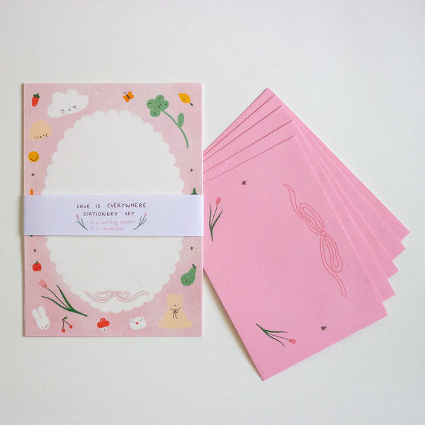 A pink stationery set displayed on a white background, featuring a scalloped white writing area, cute illustrations, and a white paper band reading 'LOVE IS EVERYWHERE STATIONERY SET', alongside a stack of matching pink envelopes.