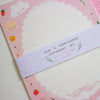 A stationery set wrapped with a white paper band that reads 'LOVE IS EVERYWHERE STATIONERY SET 10 x writing papers 5 x envelopes'. The papers are light pink with scalloped edges and cute illustrations of strawberries, smiling faces, and clouds.