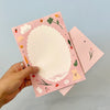 A hand holds a pink paper with a scalloped white writing area and cute illustrated border, with a matching pink envelope behind it on a light blue background.