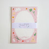 A stationery set in a clear plastic sleeve, featuring a pink cover decorated with cute illustrations of a bunny, strawberry, clover, and clouds around a large white scalloped oval. A white paper band wrapped around the center reads 'LOVE IS EVERYWHERE STATIONERY SET'.