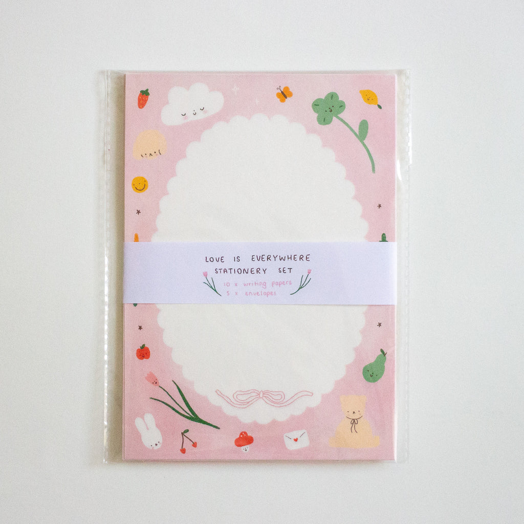 A stationery set in a clear plastic sleeve, featuring a pink cover decorated with cute illustrations of a bunny, strawberry, clover, and clouds around a large white scalloped oval. A white paper band wrapped around the center reads 'LOVE IS EVERYWHERE STATIONERY SET'.