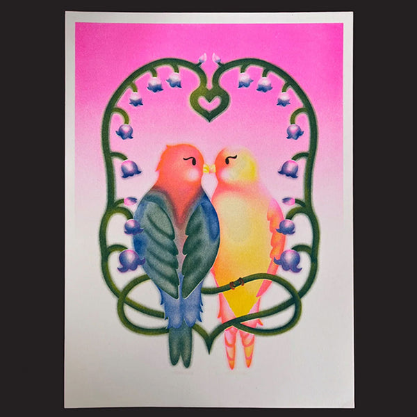 A colourful art print featuring two kissing birds framed by a heart-shaped floral border, displayed on a white sheet against a dark background.