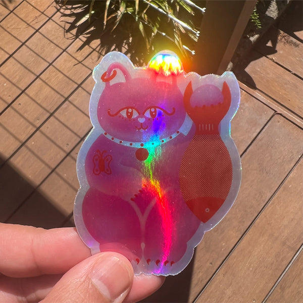 A hand holds a die-cut holographic sticker featuring a pink cartoon cat holding a fish, photographed outdoors on a wooden deck with sunlight.