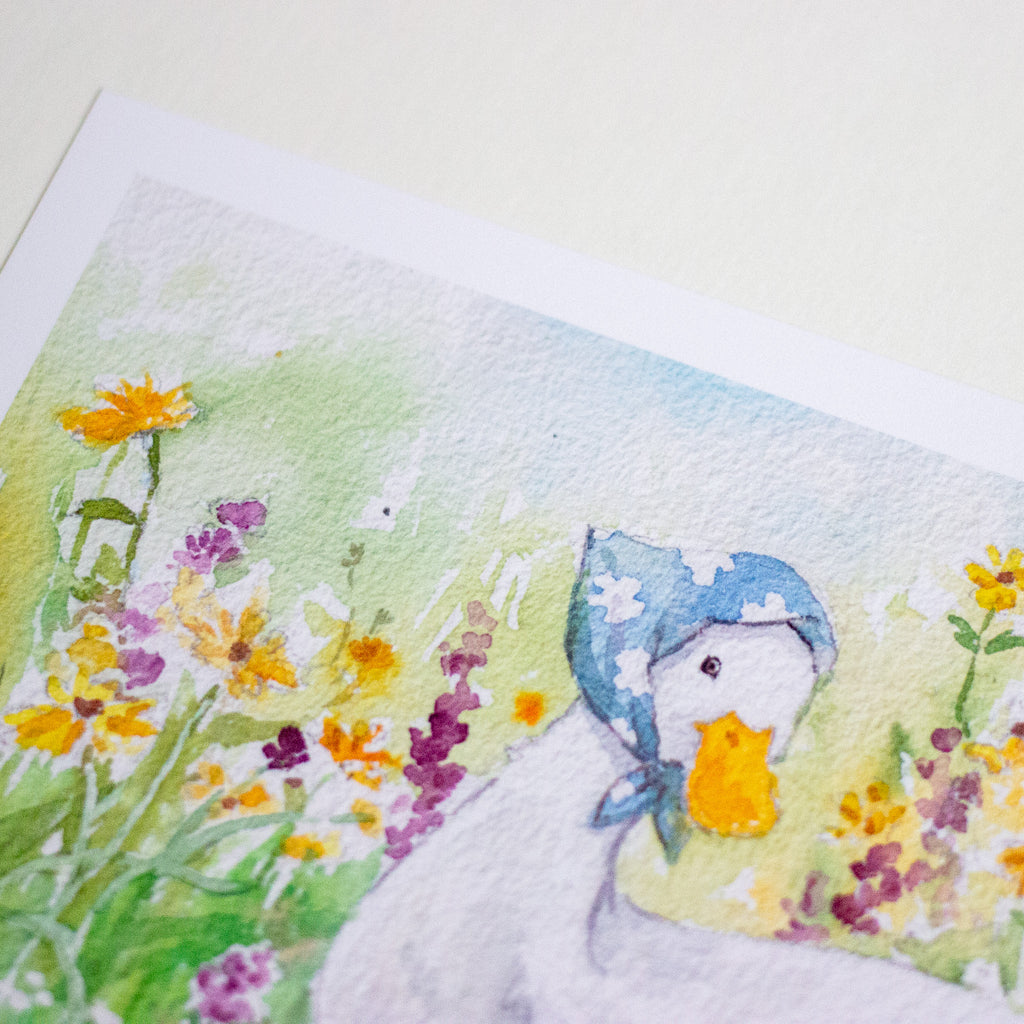 A close-up of a watercolour art print depicting a white duckling wearing a blue bonnet and standing in a field of yellow, orange, and purple wildflowers. The print is shown at an angle on a light cream-coloured surface.