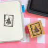 A small square wooden stamp with a black printed stamp design of a girl in a plaid dress, shown next to a notebook and an ink pad on a pink and white checkered surface.