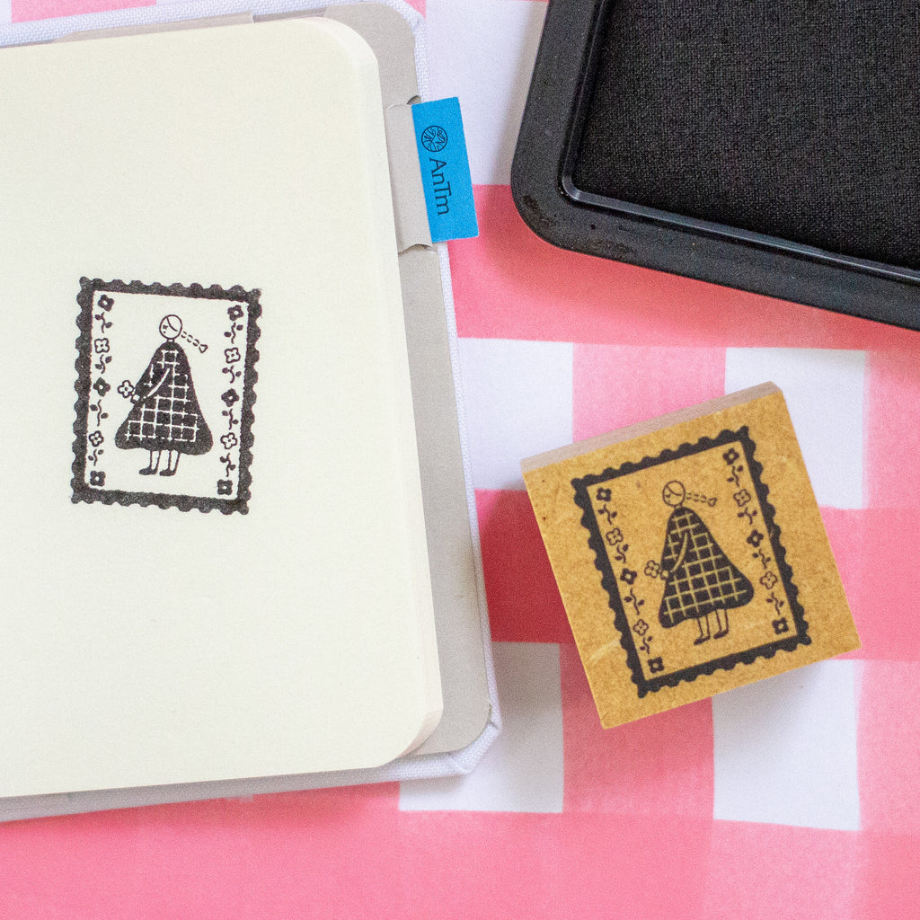 A small square wooden stamp with a black printed stamp design of a girl in a plaid dress, shown next to a notebook and an ink pad on a pink and white checkered surface.