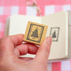 A hand holds a small square wooden stamp with a black printed design, with an open notebook and pink-and-white checkered surface blurred in the background.