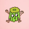 A cute embroidered patch of a smiling green canele character with pink arms and legs, set against a solid pink background.