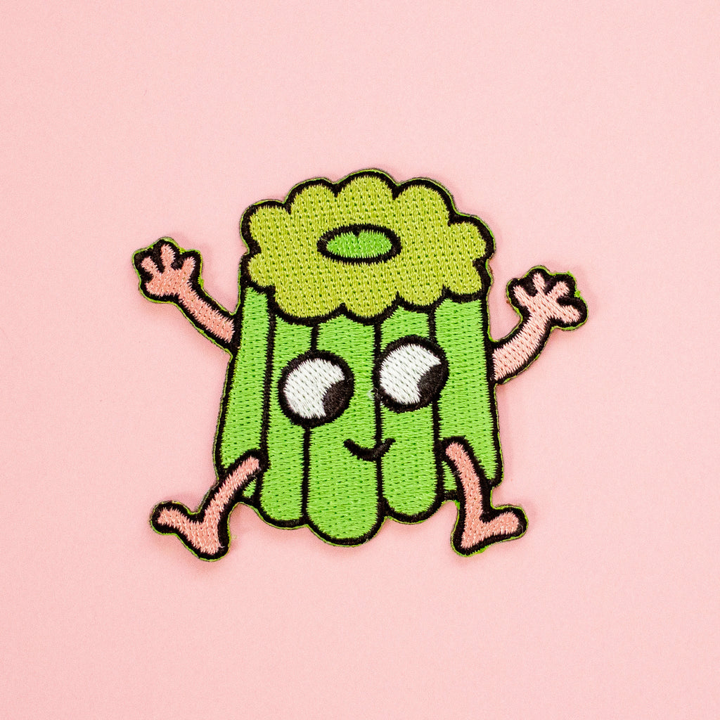 A cute embroidered patch of a smiling green canele character with pink arms and legs, set against a solid pink background.