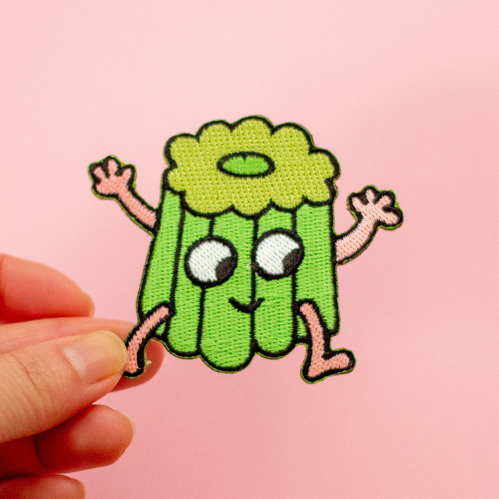A small embroidered patch shaped like a smiling green canele character with pink arms and legs, held by a person's fingers against a soft pink background.