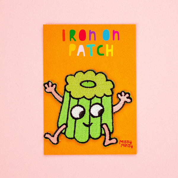 An embroidered patch featuring a green canele character with pink arms and legs, set against a bright orange background with the text 'IRON ON PATCH' in multicoloured letters. The patch is signed 'Missy Minzy' in the bottom right corner and is displayed on a light pink background.