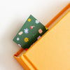 A close-up of a dark green paper bookmark with a colourful floral pattern, tucked between an orange textured notebook and a white page.