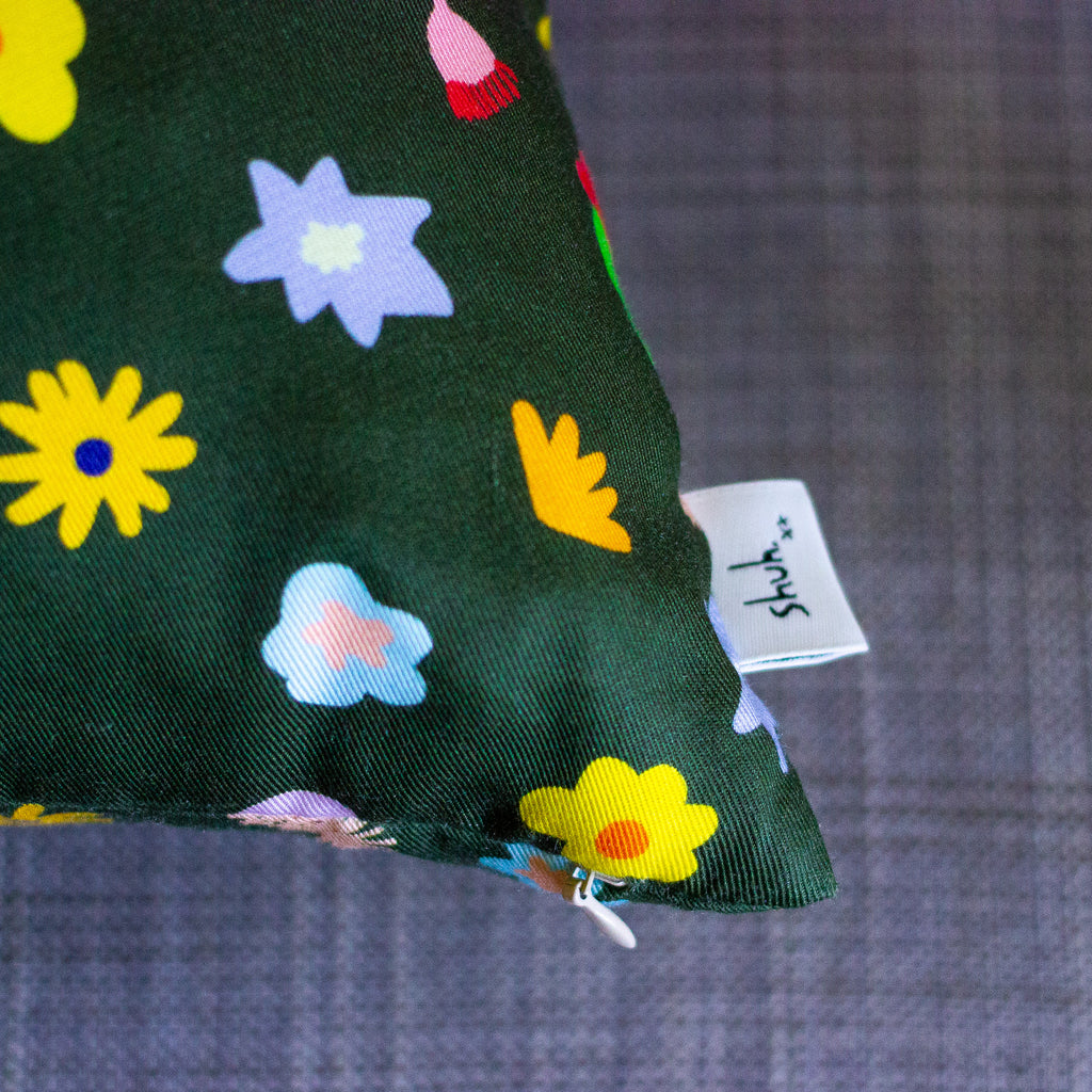A picture of the meadow cushion cover on a couch, focusing on the shuh tag and zipper. Multicoloured and varied flowers on a dark green background. 