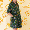A front view of the Meadow linen dress, models hands are in the pockets. With multicoloured flower designs on a meadow green background. 