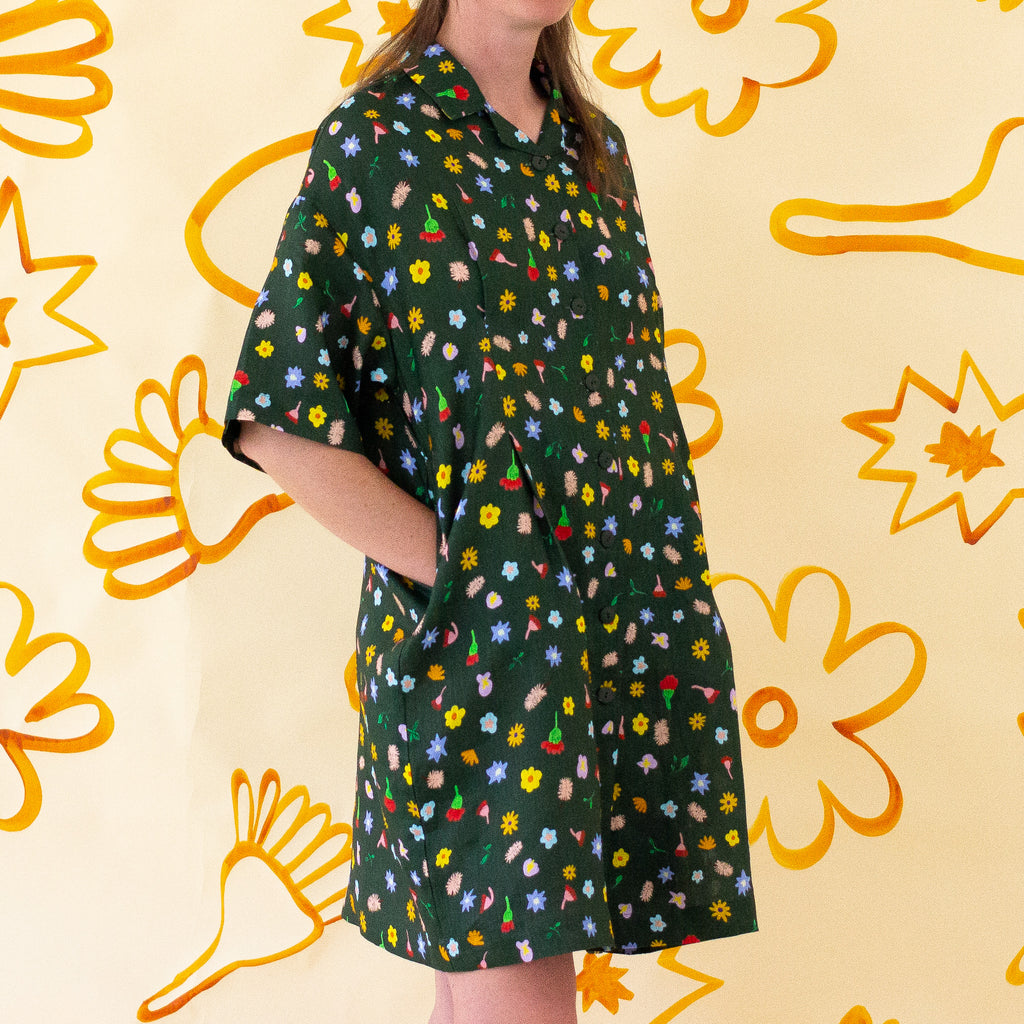 A front view of the Meadow linen dress, models hands are in the pockets. With multicoloured flower designs on a meadow green background. 