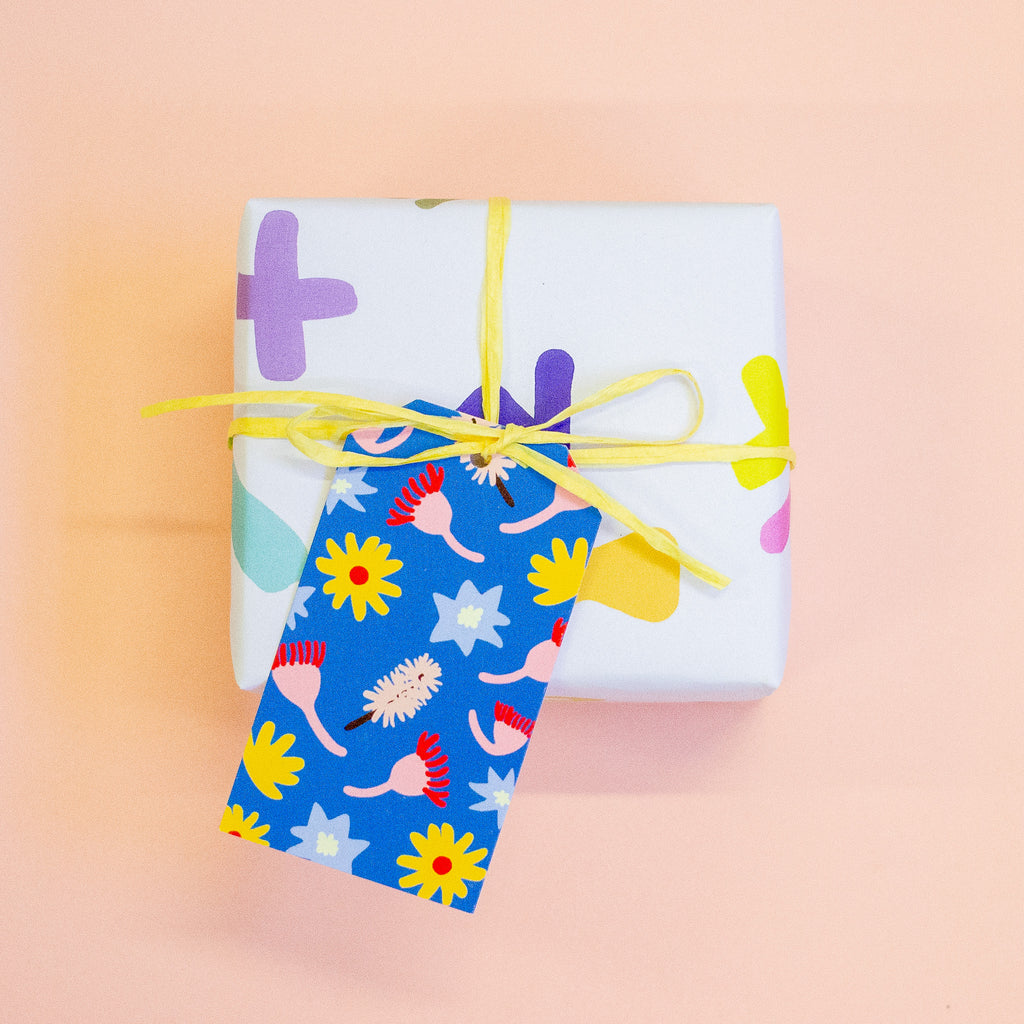 A gift tag with colourful floral and abstract patterns on a blue background attached to a yellow string on a gift box.