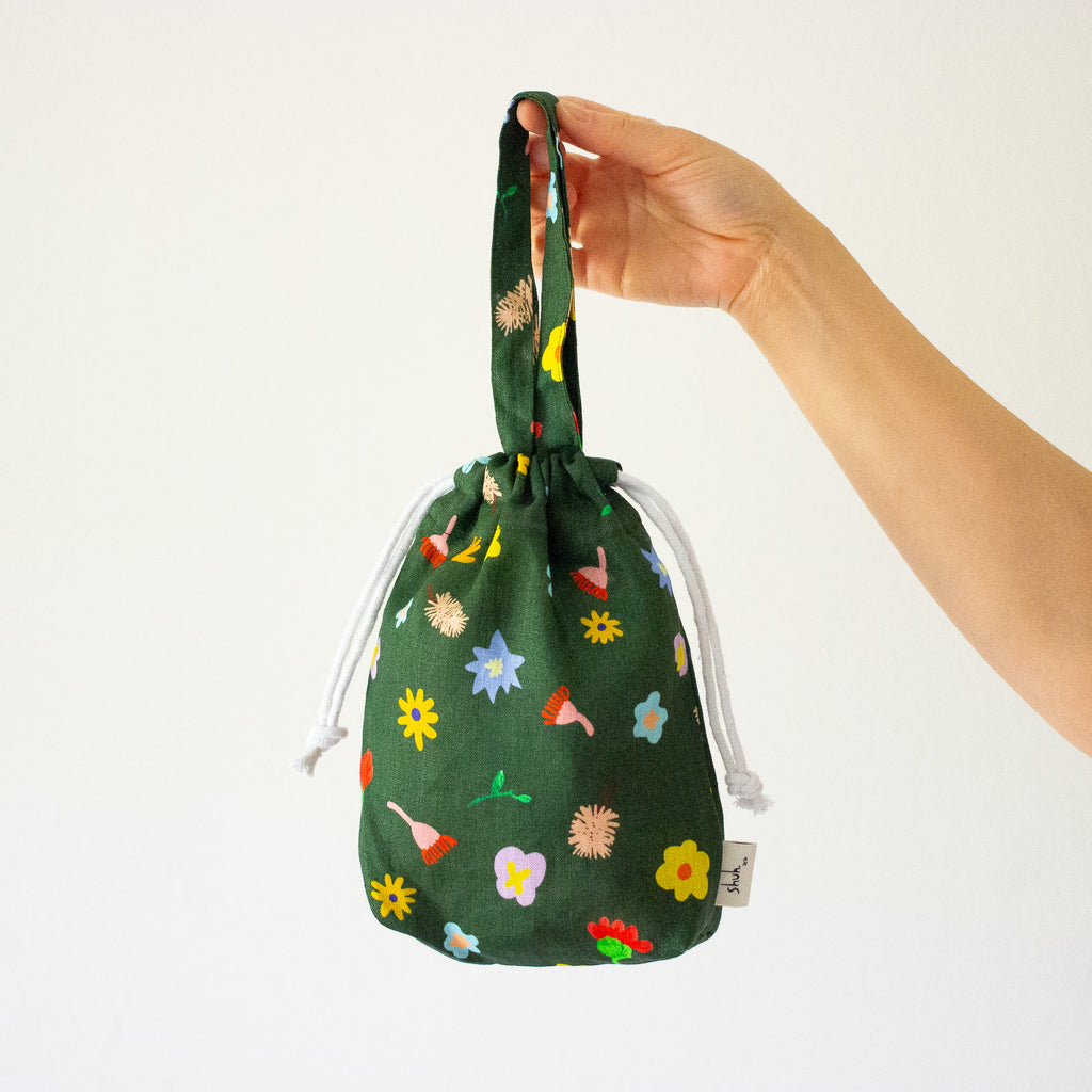 Front view of a hand holding the mini carry bag. Bag features Illustrated wildflowers on a meadow green background.