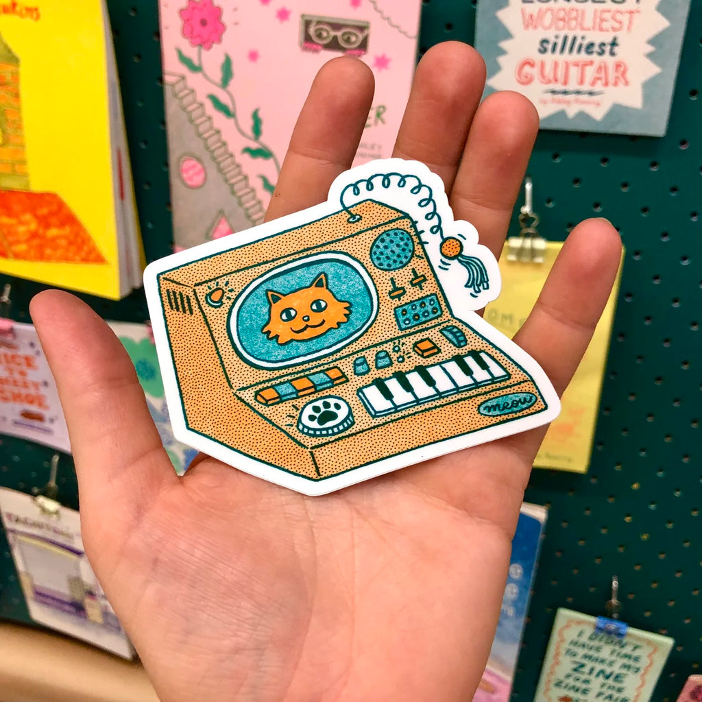 A hand holding a die-cut sticker featuring a retro, dotted-style illustration of a cat connected to a toy synthesizer. The cat is depicted inside a television screen on the synthesizer, and a coiled toy tail with a tassel hangs from the top right.