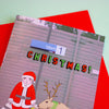 A Christmas-themed greeting card featuring a station platform scene with the text 'Merri CHRISTMAS!' and an illustration of Santa Claus and a reindeer, layered over a bright red envelope on a pale mint background.