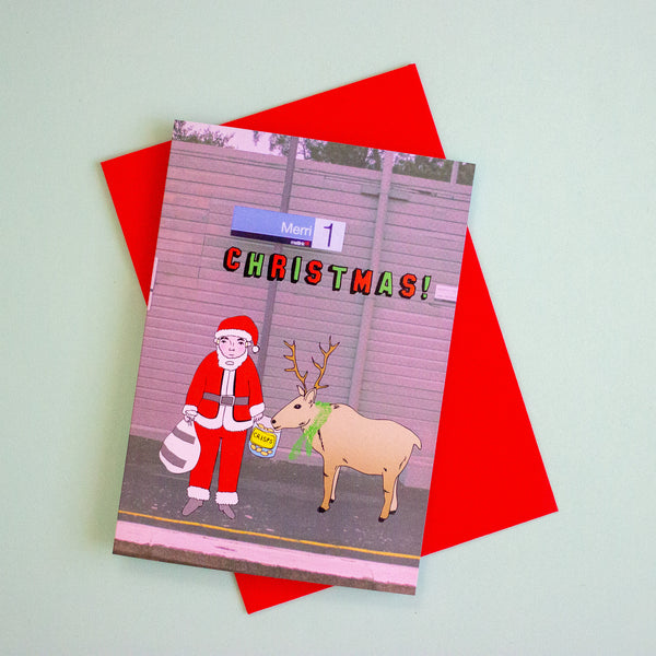 A Christmas greeting card featuring a cartoon Santa Claus and a reindeer in front of a grey wall with the word 'CHRISTMAS!' in colourful letters, shown on top of a bright red envelope against a pale mint background.