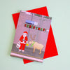 A Christmas greeting card featuring a cartoon illustration of Santa Claus feeding a reindeer, layered over a bright red envelope on a pale mint green background.