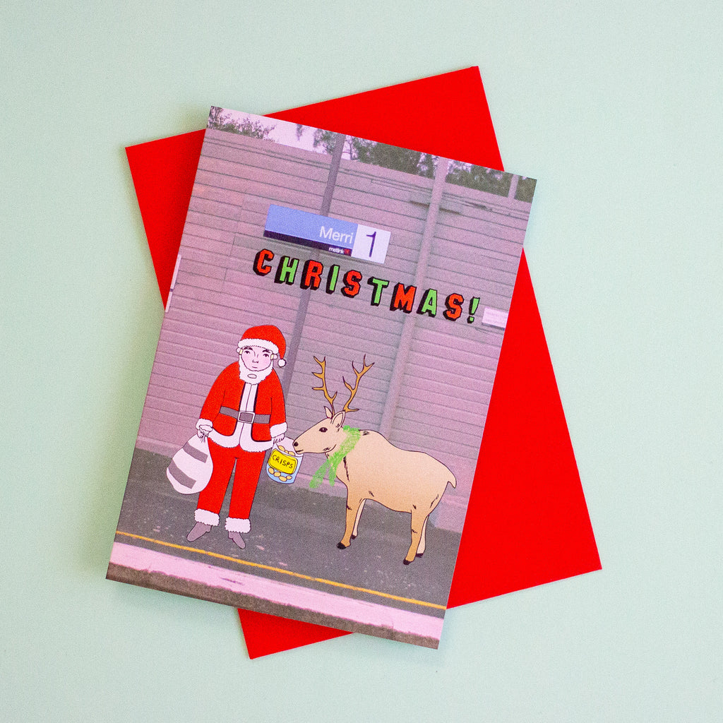 A Christmas greeting card featuring a cartoon illustration of Santa Claus feeding a reindeer, layered over a bright red envelope on a pale mint green background.
