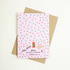 A pink greeting card with a red and green polka dot pattern and small illustrations near the bottom, placed on top of a brown kraft envelope against a light cream background. The card reads 'shuh.