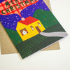 A Christmas greeting card featuring a stylised yellow house with a red roof, green hills, and a dark blue night sky with stars, placed on top of a brown envelope against a white background.