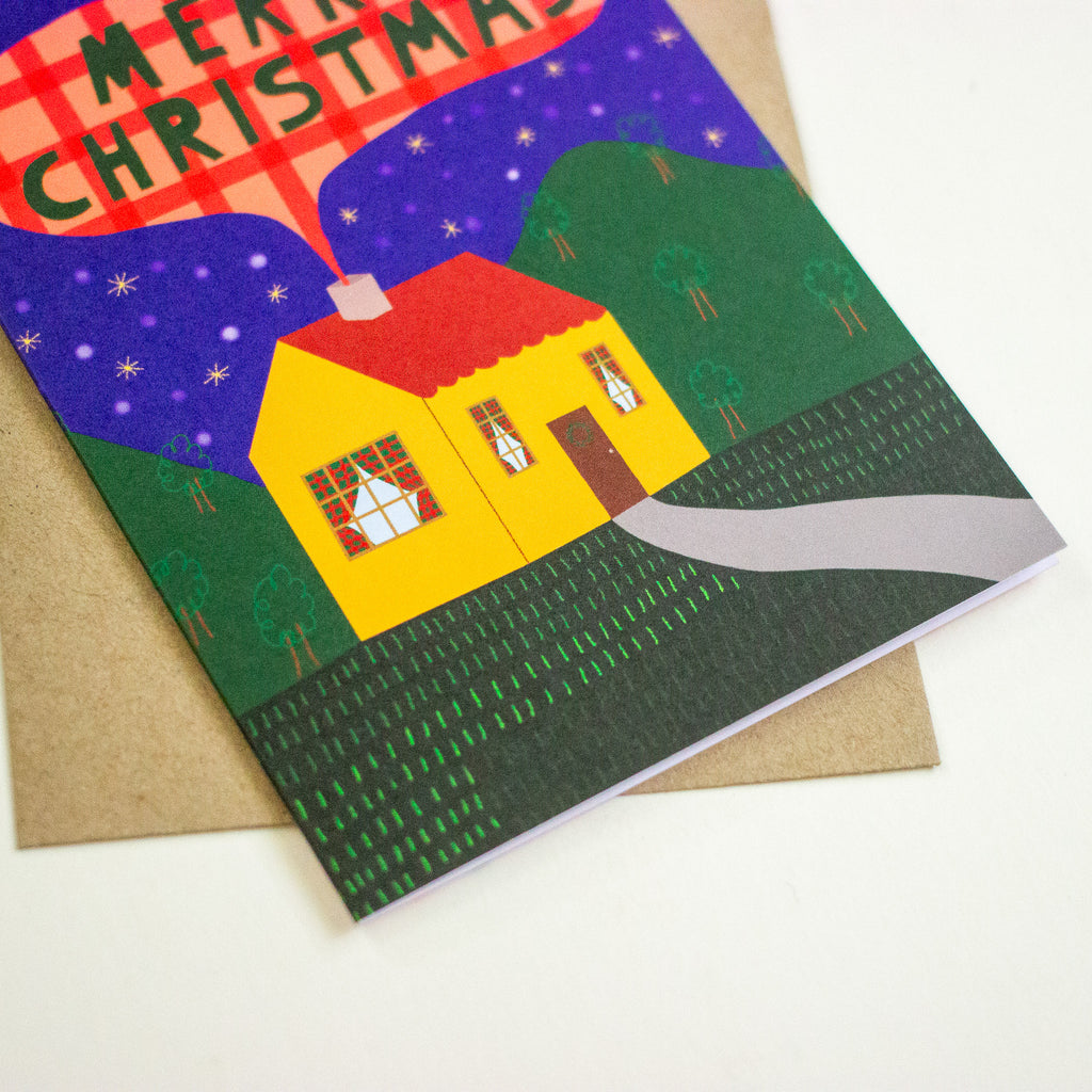 A Christmas greeting card featuring a stylised yellow house with a red roof, green hills, and a dark blue night sky with stars, placed on top of a brown envelope against a white background.
