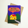 A Christmas greeting card featuring a stylised yellow house with a red roof, green hills, and a dark blue night sky with stars, placed on top of a brown envelope against a white background.