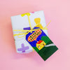 A Christmas-themed gift tag featuring a hand-painted style illustration of a yellow house, green hills, and a blue starry night sky with a red plaid banner reading 'MERRY CHRISTMAS' attached to a yellow string on a gift box. 
