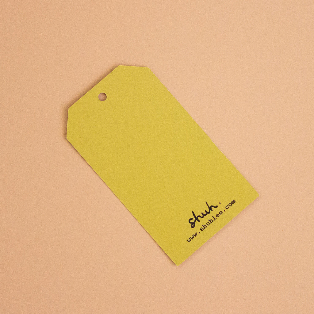 A yellow paper hang tag with a hole at the top left, printed with the brand name 'shuh.' and website 'www.shuh.com'
