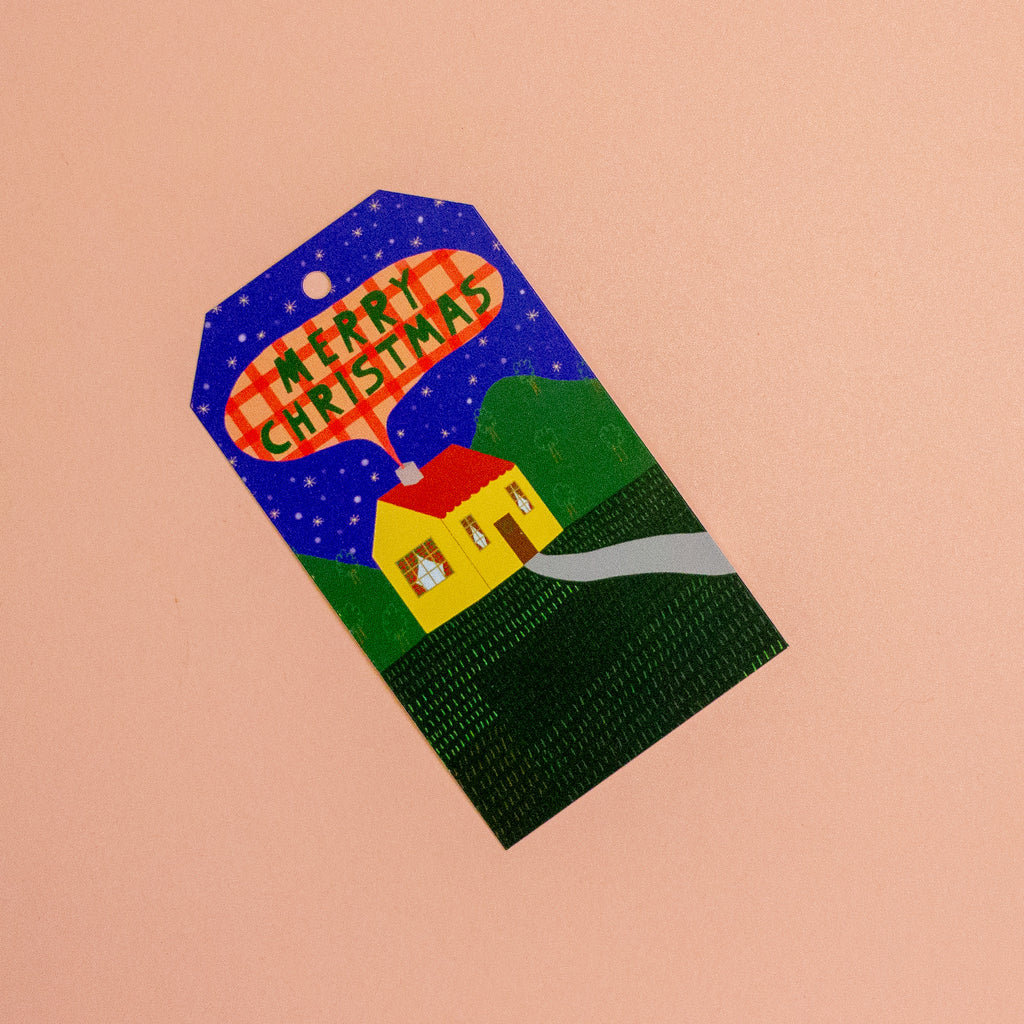 A Christmas-themed gift tag featuring a hand-painted style illustration of a yellow house, green hills, and a blue starry night sky with a red plaid banner reading 'MERRY CHRISTMAS'.
